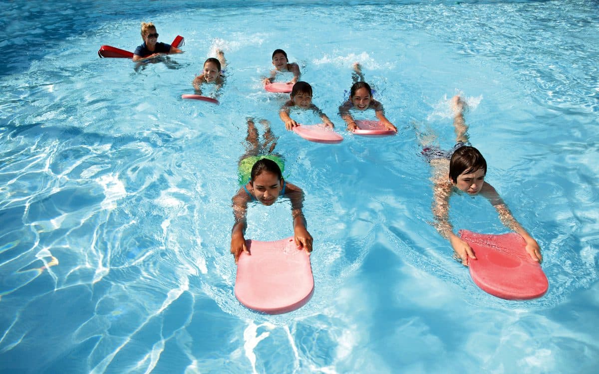 Children practising kicking in a pool with kickboards while an instructor watches